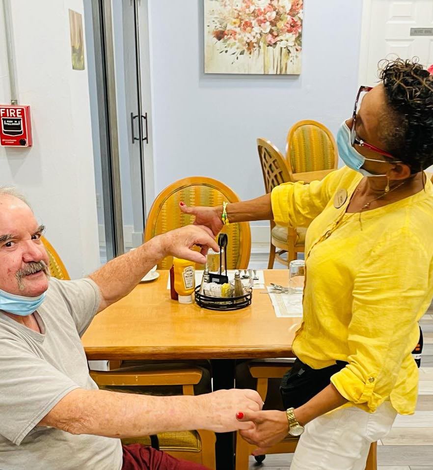A healthcare worker assists an elderly man at a care facility, both wearing masks.