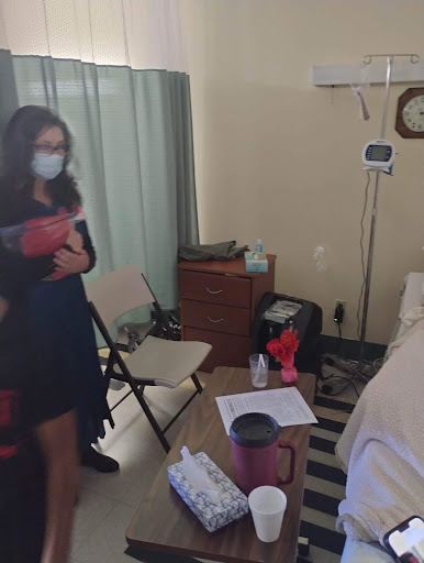 A caregiver in a mask interacts with a patient in a hospital room.
