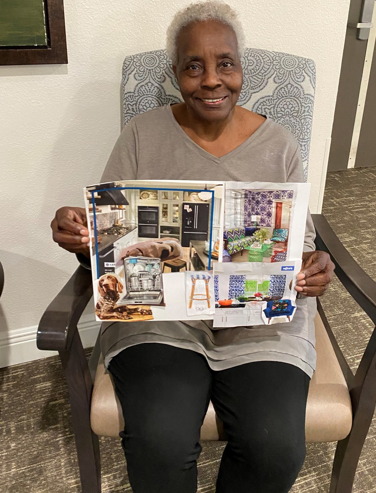 An elderly woman seated indoors, holding up a collage artwork.