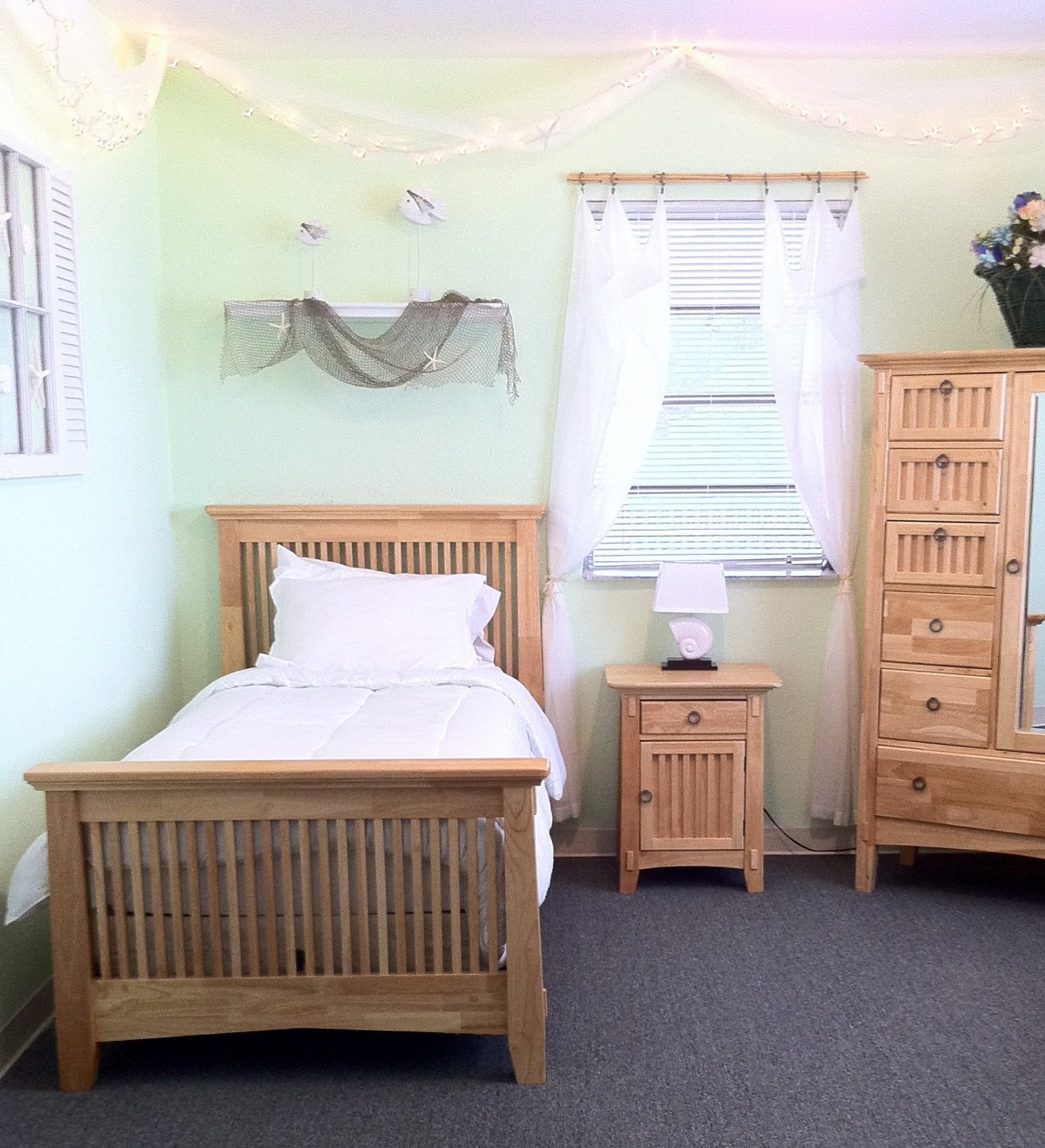 A cozy bedroom with light wooden furniture, including a single bed, nightstand, and dresser against light green walls.