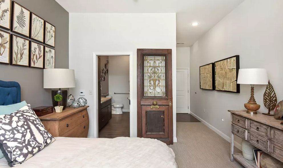 A modern bedroom featuring a cozy bed, wooden dresser, and an elegant lamp, with a glimpse into an adjoining bathroom.