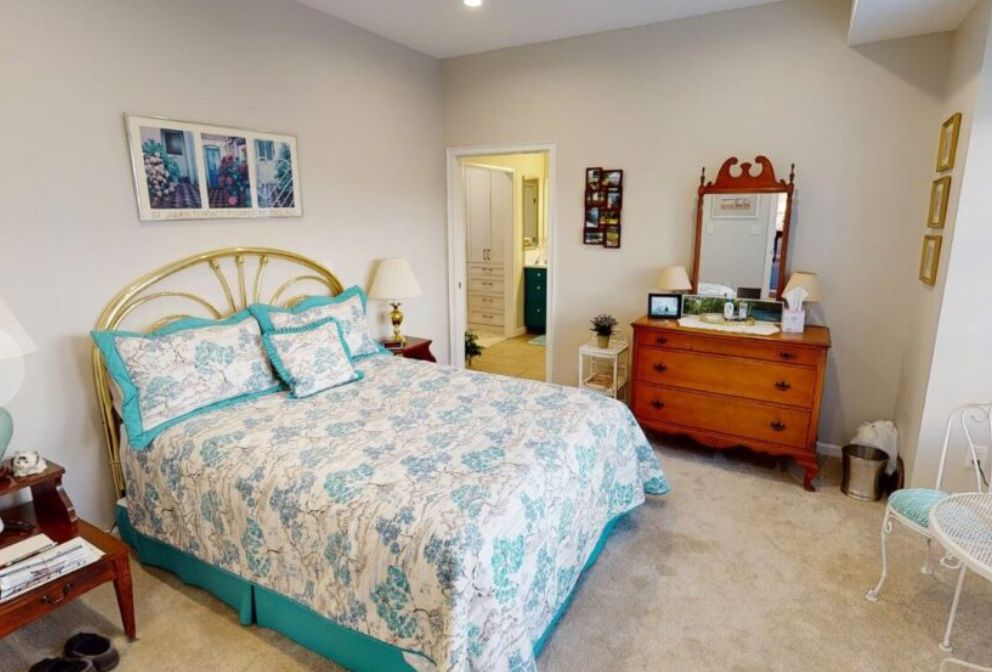 A cozy bedroom featuring a neatly made bed with patterned bedding, a wooden dresser with a mirror, and soft carpeting.