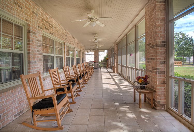 A spacious porch with several wooden rocking chairs lined up against a brick wall, featuring ceiling fans and bright natural light.