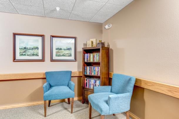 A cozy waiting area featuring two blue armchairs, a bookshelf filled with books, and framed landscape paintings on the wall.