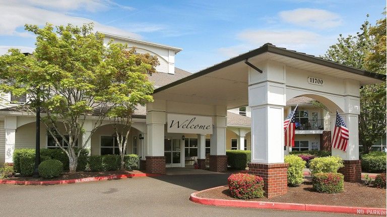 The image shows the entrance of a building with a 'Welcome' sign, surrounded by well-maintained landscaping.