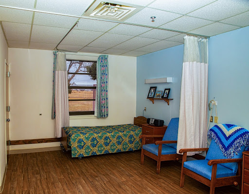 A quiet hospital room featuring a bed, two chairs, and blue walls.