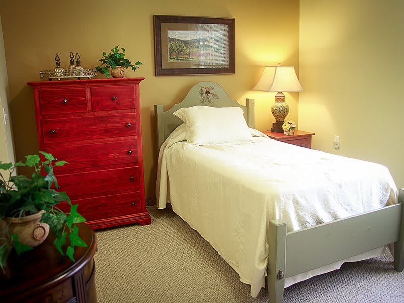 A cozy bedroom with a single bed, red dresser, and soft lighting from a table lamp.