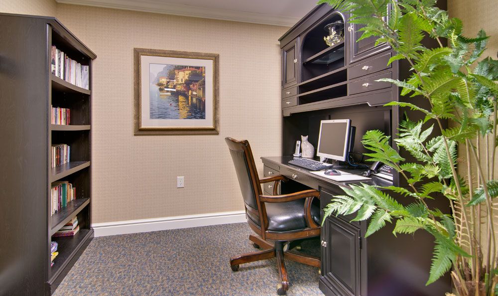 A cozy home office with a wooden desk, computer, and comfortable chair, accented by plants and artwork on the wall.