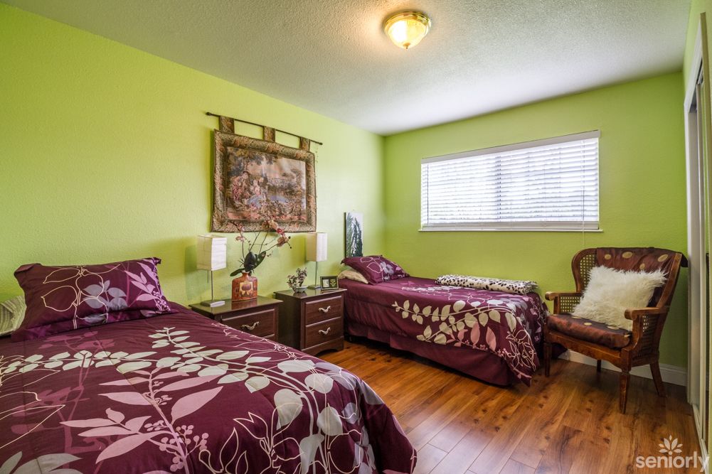A cozy bedroom featuring twin beds with purple floral bedding, set against vibrant green walls and a wooden floor. A decorative tapestry hangs above the nightstand, and natural light filters in through a window with blinds.