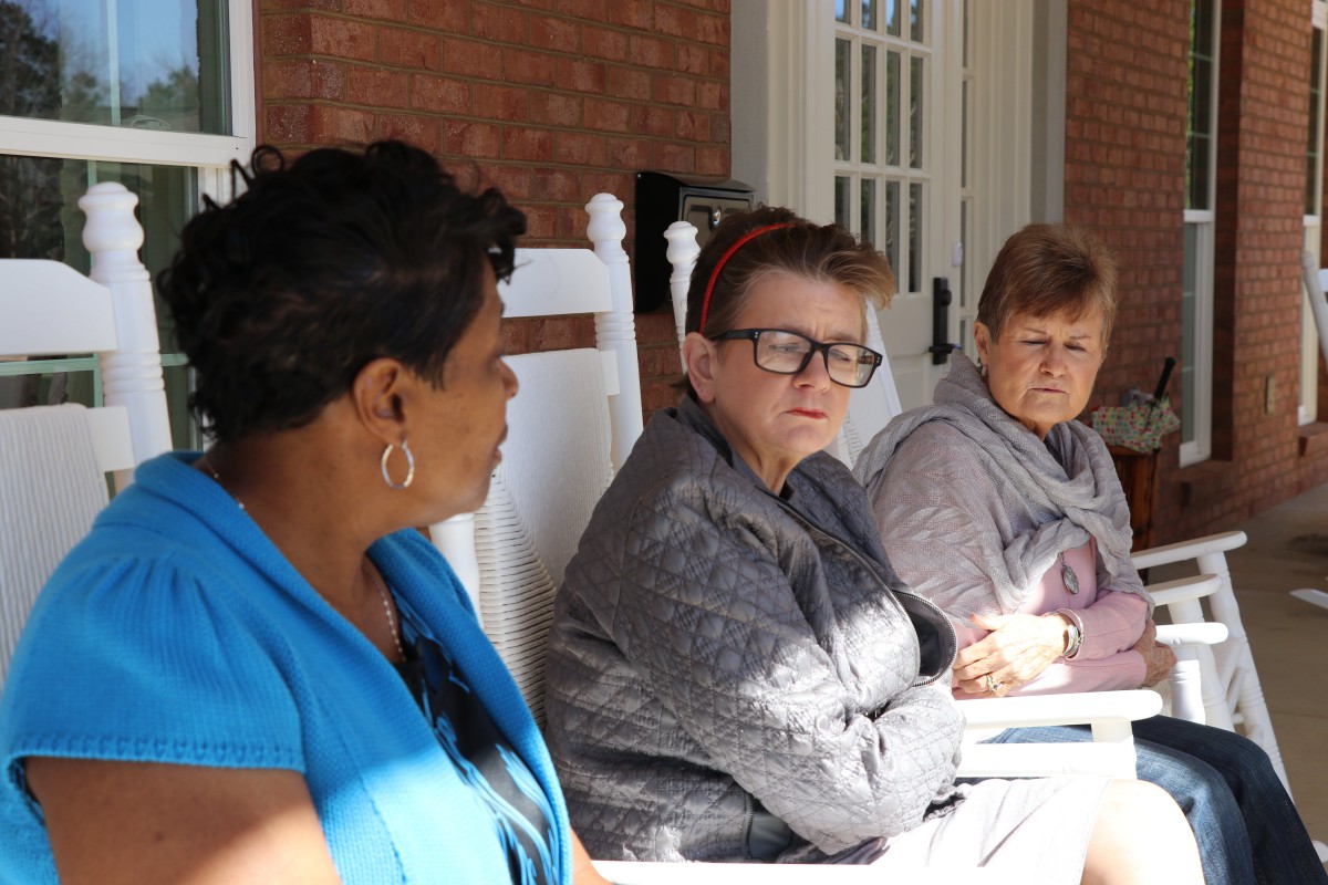 Three people sitting and conversing on a porch during the daytime.