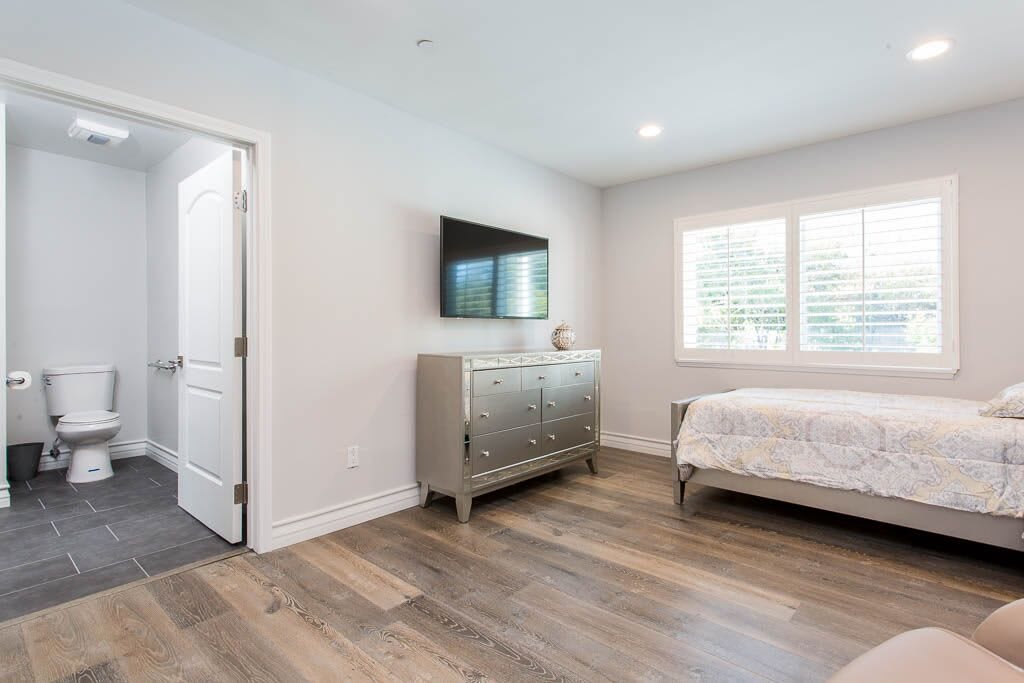 A modern, minimalist bedroom with a hardwood floor, featuring a bed, dresser, and mounted TV.