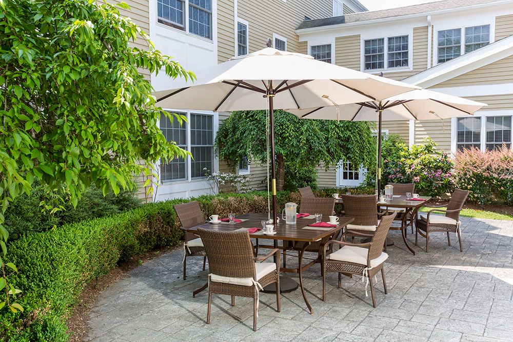 A cozy outdoor patio area with a table and chairs under a large umbrella, surrounded by greenery and the exterior of a building.