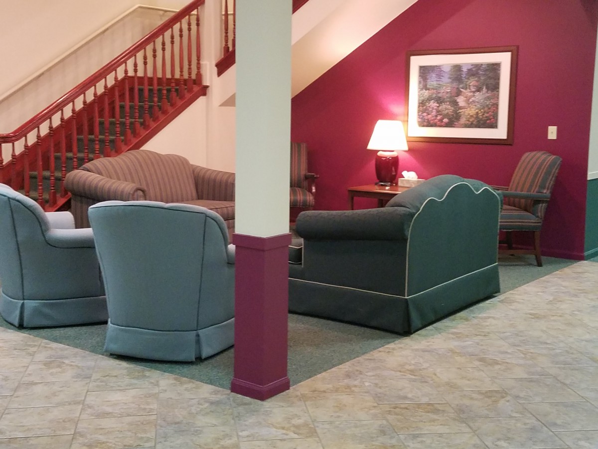 A cozy lobby area with sofas and an armchair arranged around a table lamp beneath a staircase.