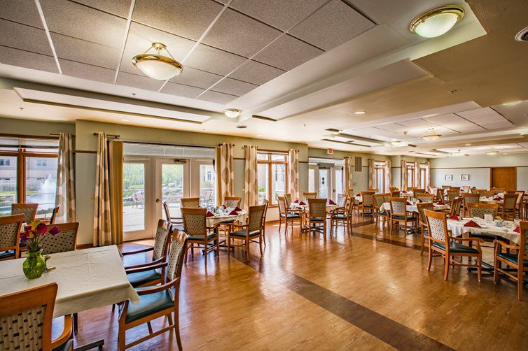 A spacious dining area with wooden floors, arranged tables and chairs, and large windows providing natural light.