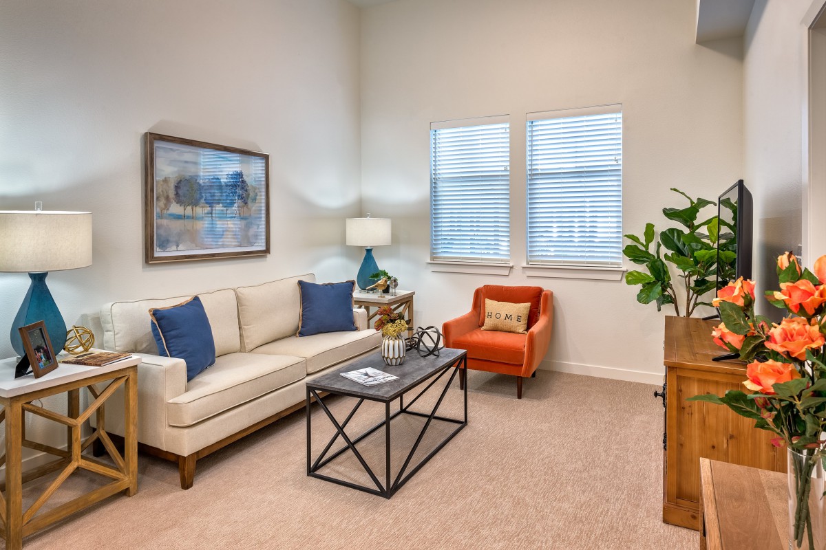 A modern living room featuring a beige sofa with blue cushions, an orange armchair, a black metal coffee table, and decorative elements such as artwork and an indoor plant.