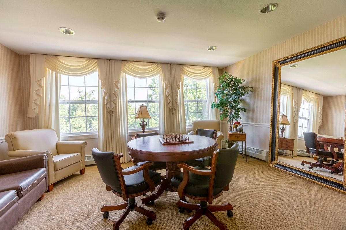 A cozy living room with a round table set for chess, surrounded by chairs and bathed in natural light from large windows.