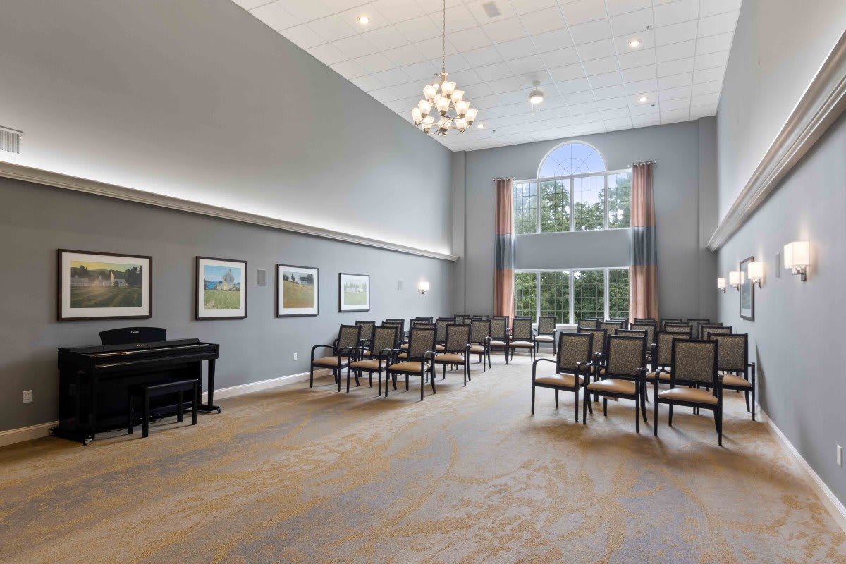 A spacious conference room with neatly arranged chairs, a piano, paintings on the walls, and a large window providing natural light.