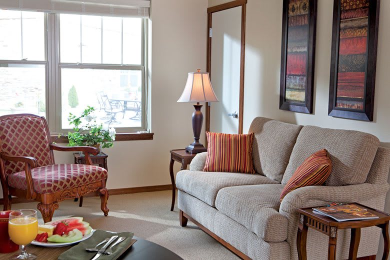 A cozy living room featuring a comfortable sofa with decorative cushions, a table lamp, and a window letting in natural light.