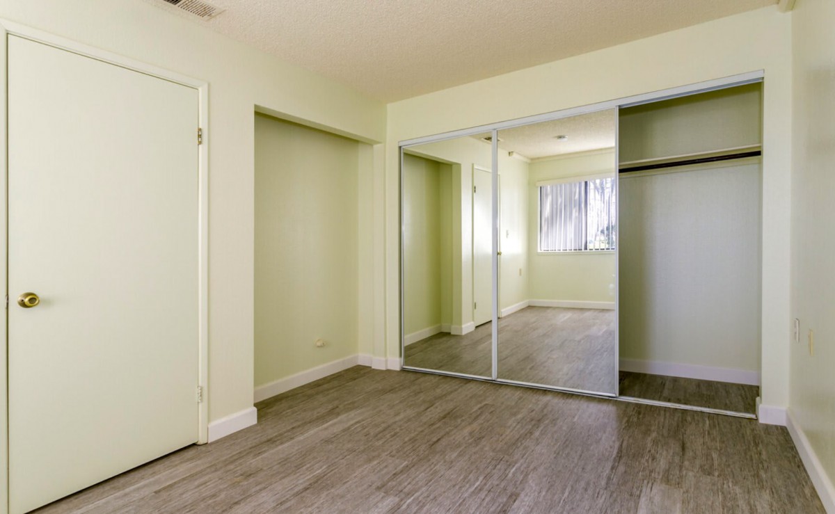 An empty room with wood flooring and mirrored closet doors, featuring light-colored walls and a window.