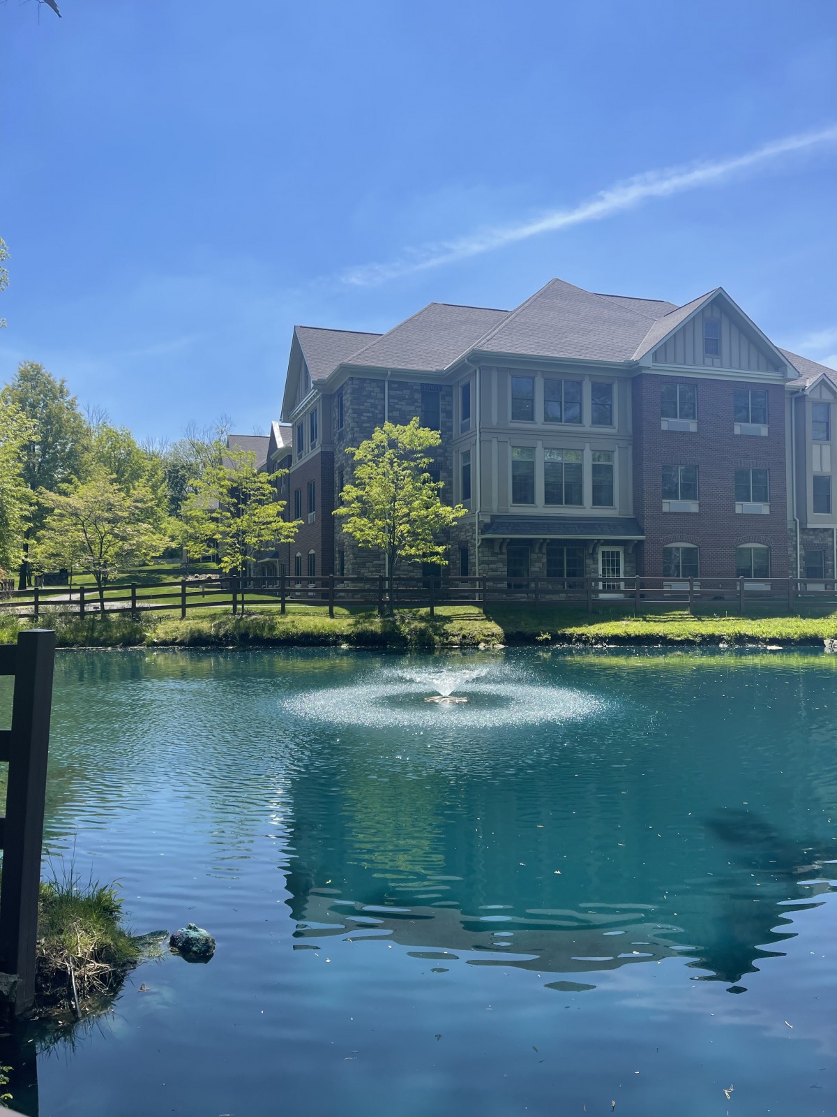 A scenic view of a building by a pond with a fountain and trees under a clear blue sky.