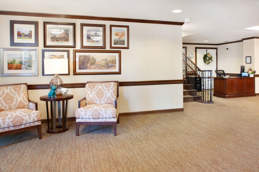A cozy interior space featuring a patterned chair, side table with a lamp, framed artwork on the wall, and a staircase in the background.