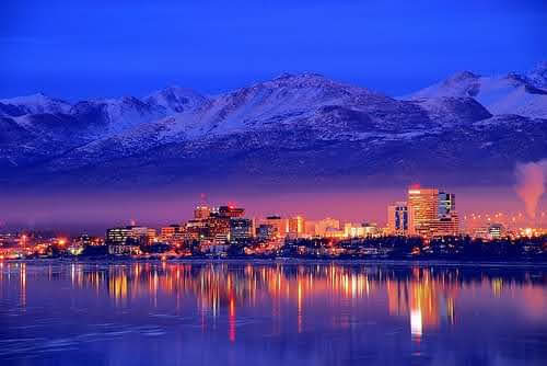 A stunning cityscape at dusk, showcasing the reflection of illuminated buildings against a serene body of water, with majestic mountains in the background.