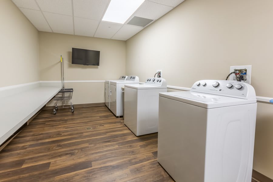 Interior of a laundromat featuring washing machines and dryers with a wooden floor and a television.
