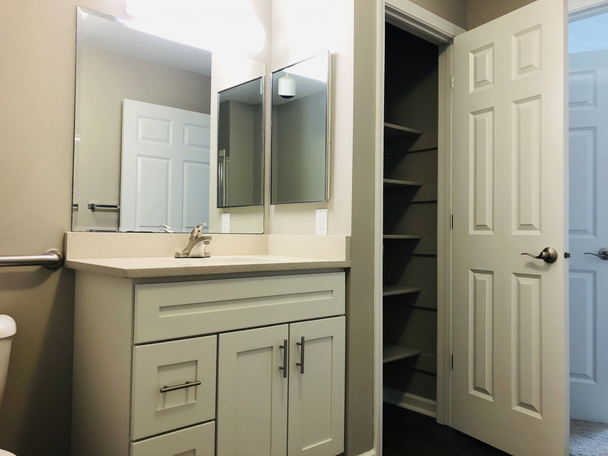 A bathroom featuring a vanity with a sink and mirror, alongside an open door revealing shelves.