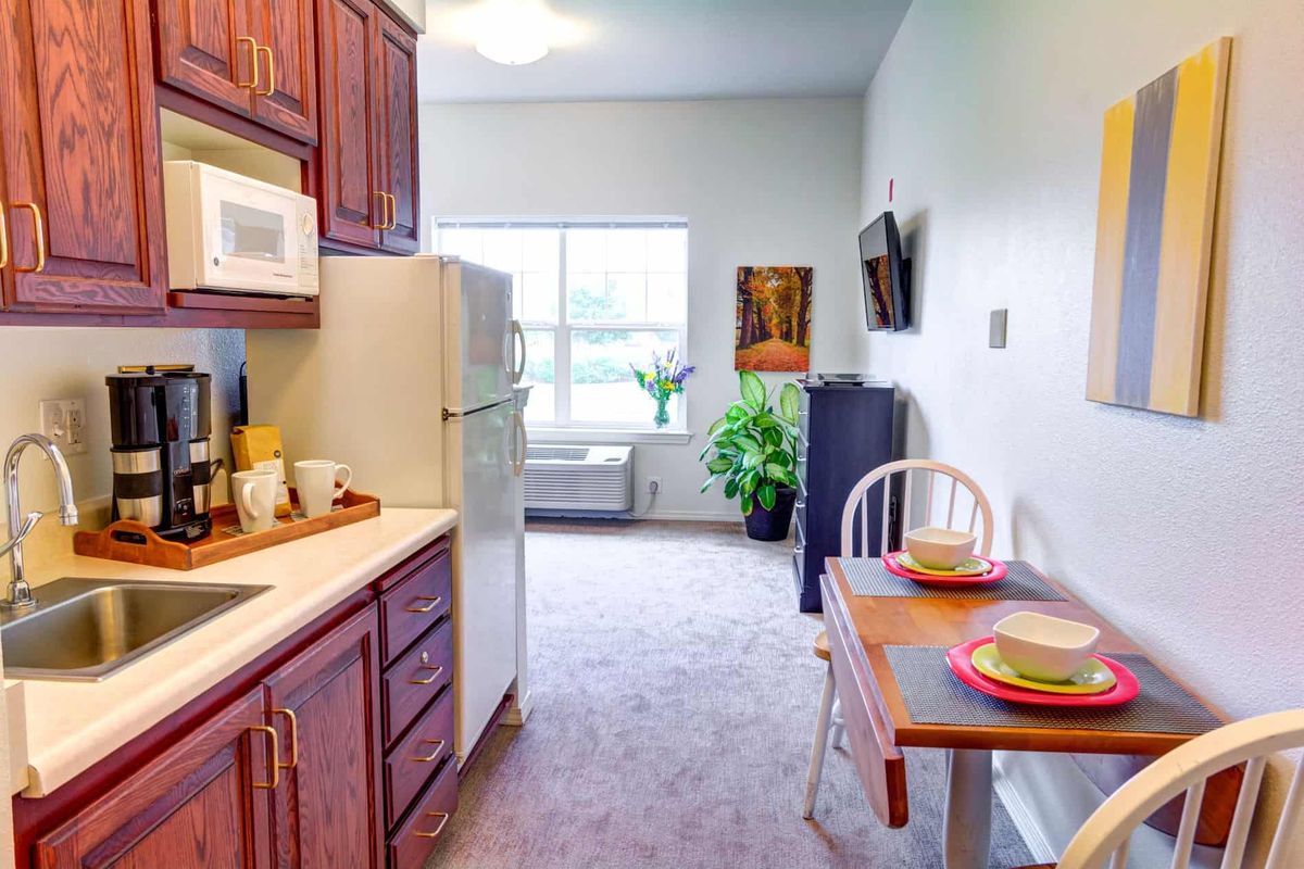 A cozy kitchen and dining area with a table set for two, featuring a refrigerator, microwave, and an indoor plant by the window.