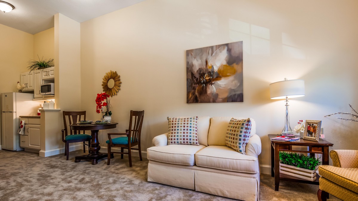 A cozy living room with a beige sofa, decorative pillows, and abstract artwork on the wall. A small table and chairs are tucked in the corner.