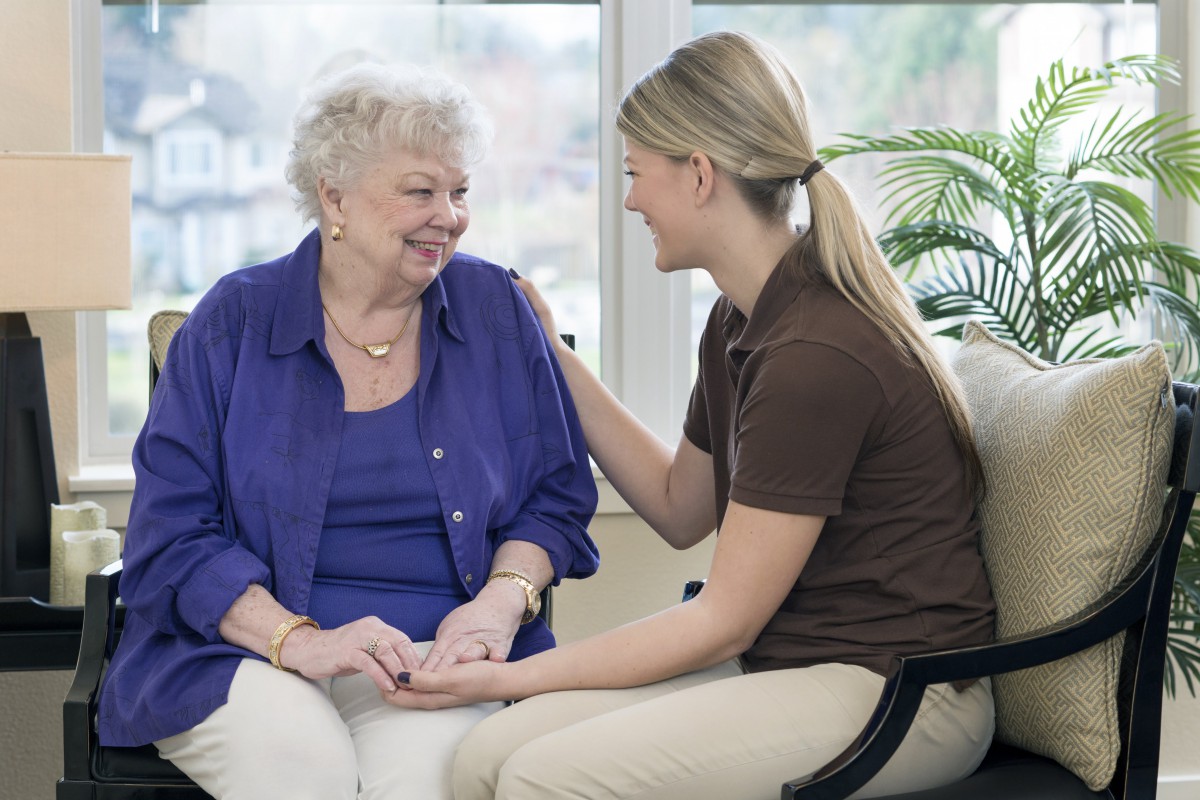 A caregiver and an elderly woman share a warm conversation indoors.