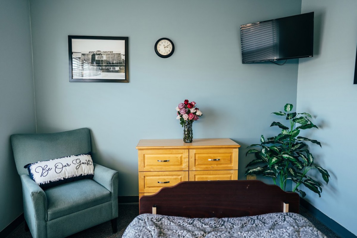 A cozy bedroom with a wooden dresser, armchair, wall-mounted TV, and a vase of flowers.