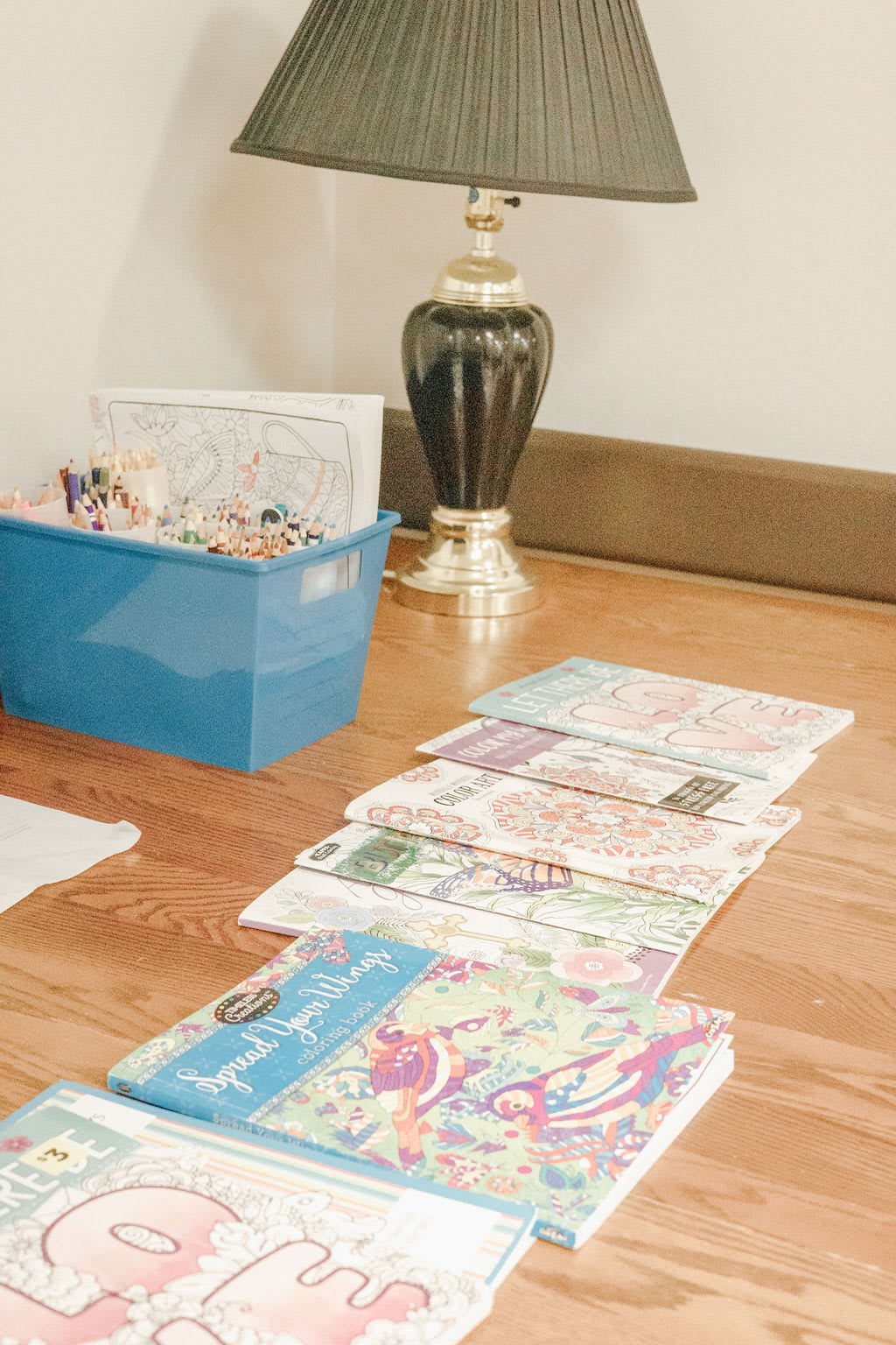 A collection of coloring books and art supplies arranged on a wooden table beside a lamp.