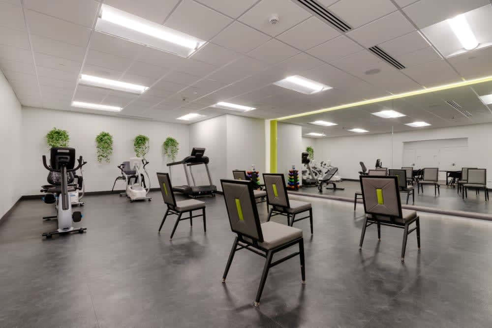 A spacious exercise room with chairs arranged in a circle, mirrors on the wall, and gym equipment in the background.