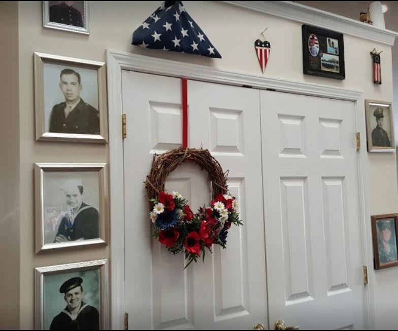 A wall decorated with framed military photographs, an American flag folded in a triangle, and a wreath hanging on double doors.