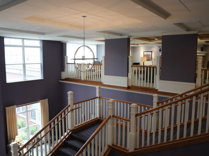 A modern building interior featuring a large staircase with wooden banisters and carpeted steps, illuminated by natural light from the windows and an overhead lighting fixture.