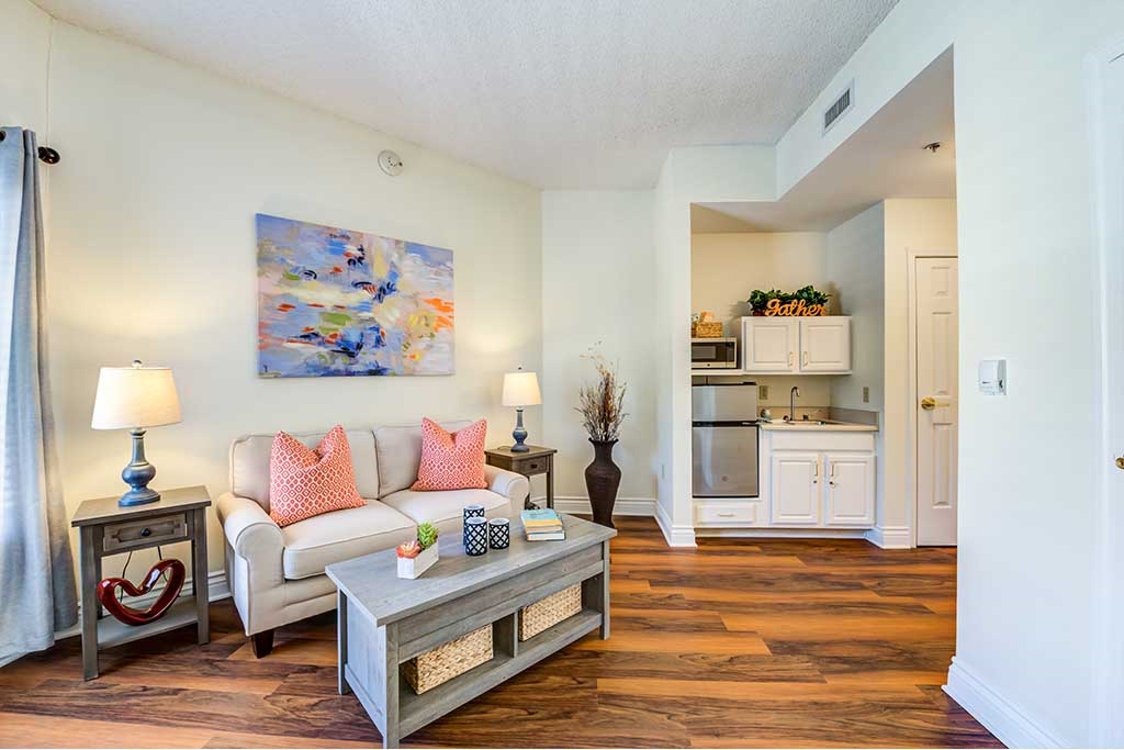 A cozy living room with a sofa, coffee table, and vibrant artwork on the wall, featuring warm wood flooring and a view into a compact kitchenette.
