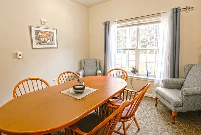 A cozy dining room with a wooden table and chairs, brightened by natural light from a large window.