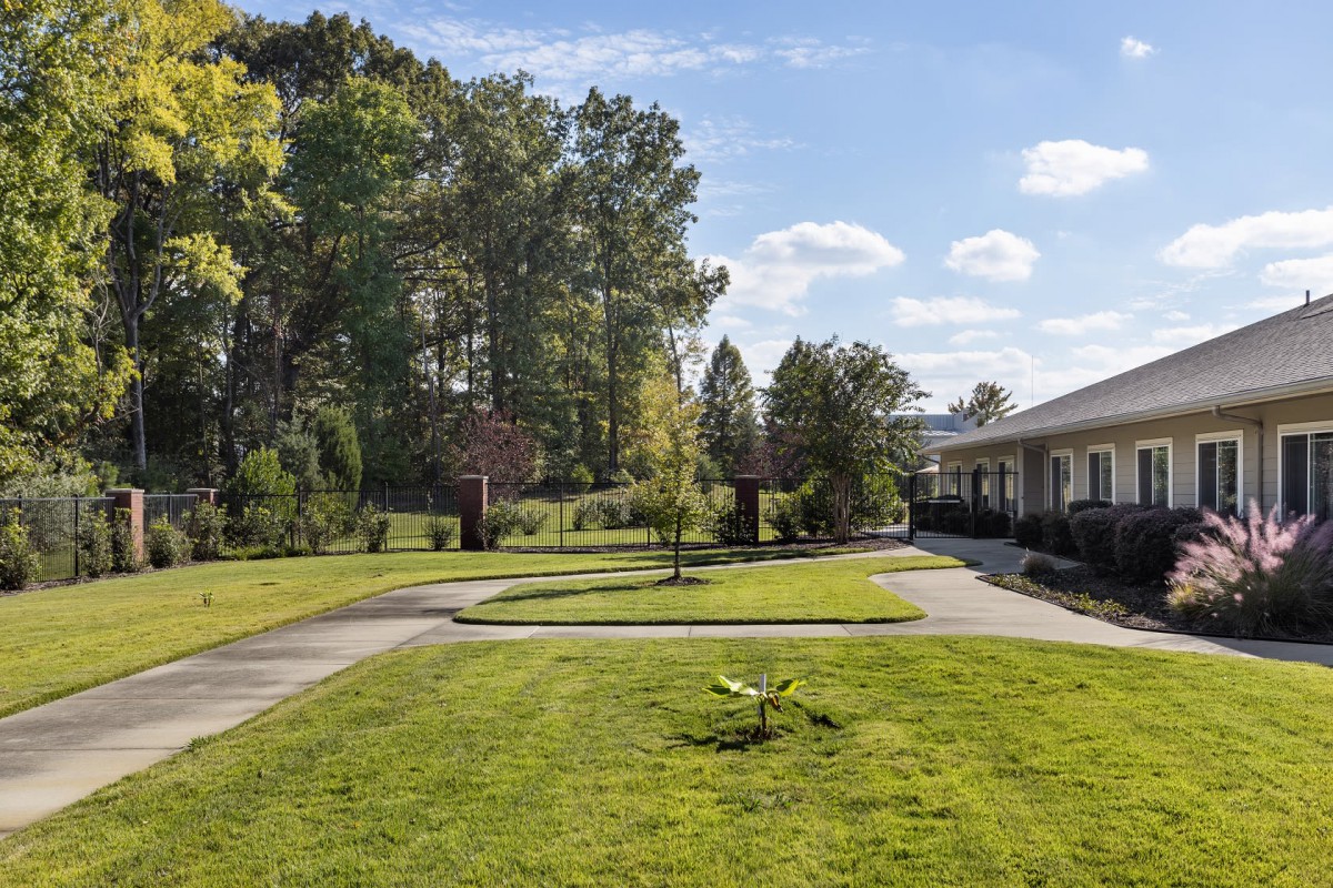 A well-maintained garden with a walkway, surrounded by trees and a building under a partly cloudy sky.