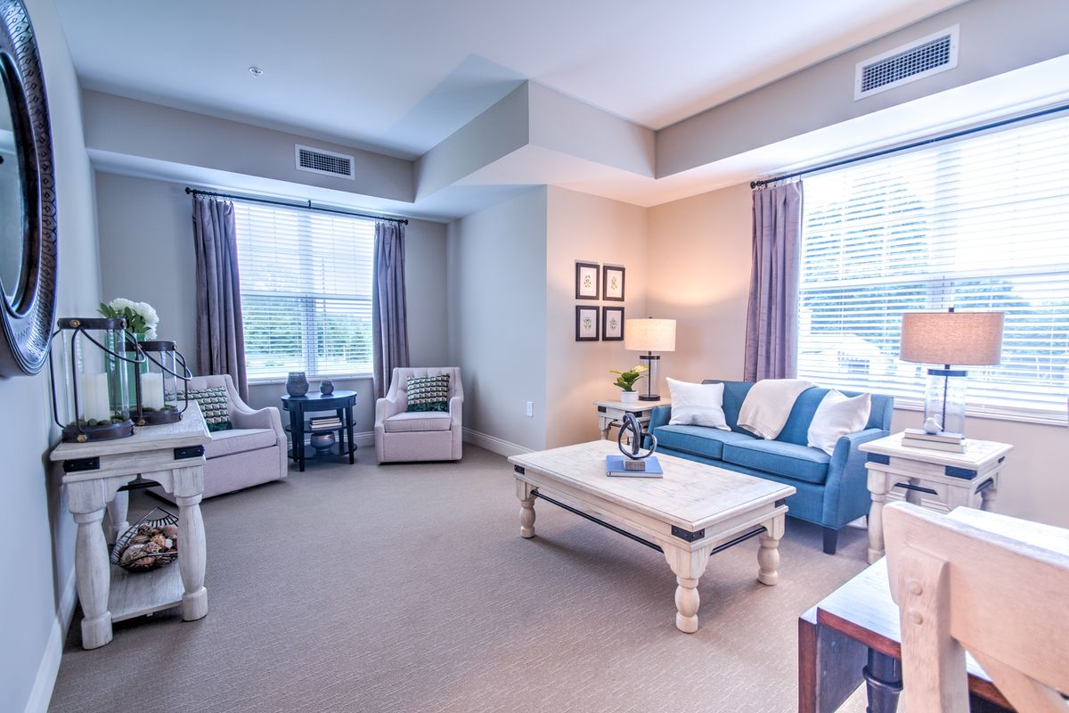 A cozy living room with a blue sofa, two armchairs, and a coffee table in the center. The room has large windows with curtains, allowing natural light to fill the space.