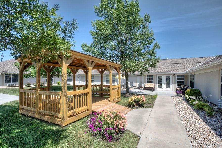 A sunny garden scene featuring a wooden gazebo, surrounded by trees and flowers with a pathway leading to a building.
