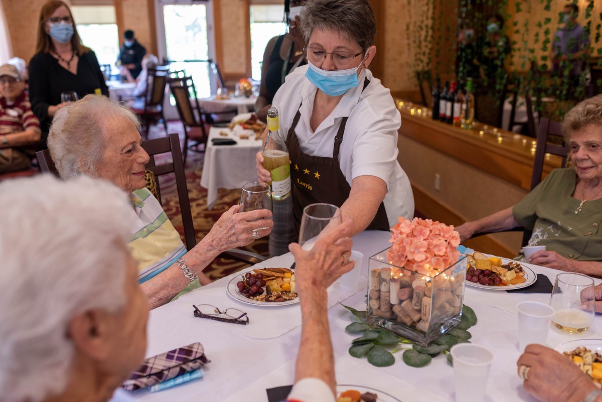 A waitress wearing a mask serves drinks to seniors at a restaurant table.