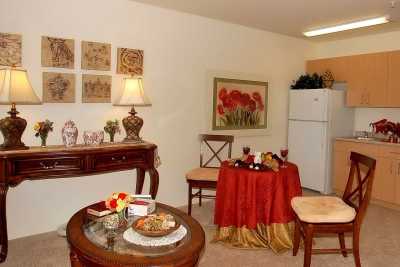 A cozy dining and living area with classic decor, featuring a small table draped in red cloth, a lamp on a side console, and a refrigerator in the background.