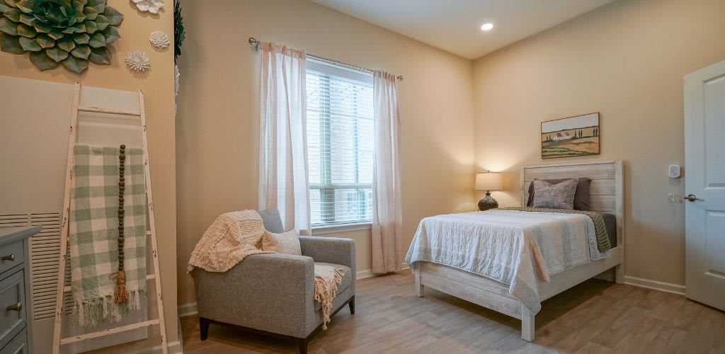 A cozy bedroom with a comfortable bed, armchair, and natural light filtering through the window.