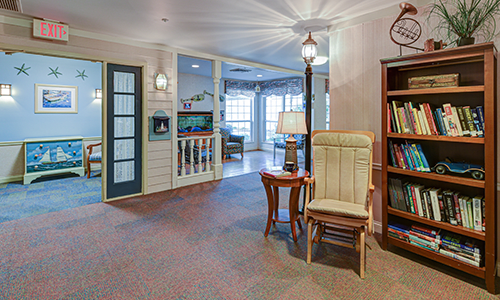 A cozy living room with a rocking chair, lamp, and bookshelf near large windows.