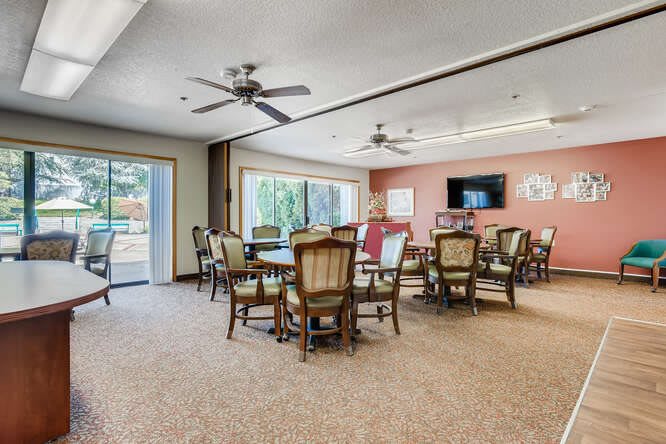 A spacious dining room with multiple tables and chairs, featuring ceiling fans, large windows, and a television on the wall.