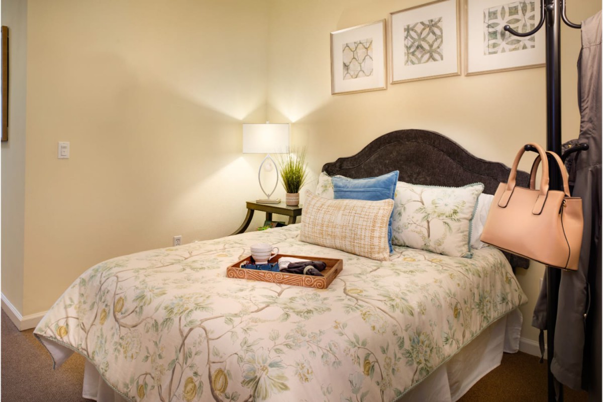 A cozy bedroom with a neatly made bed, featuring floral bedding and decorative pillows. The room is softly lit by a table lamp on the nightstand, and framed artwork adorns the wall.