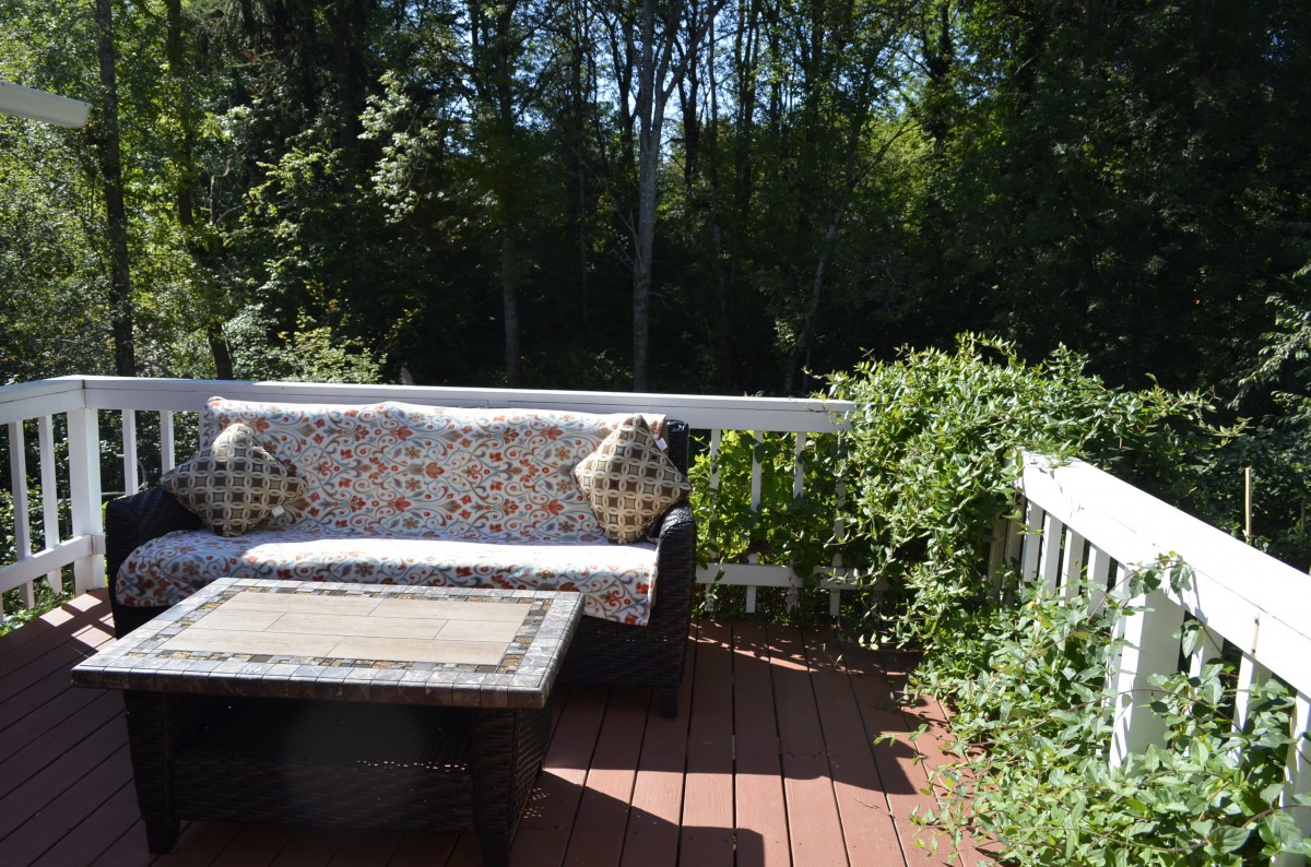 A cozy outdoor patio with a sofa, cushions, and a table set on a wooden deck surrounded by greenery.