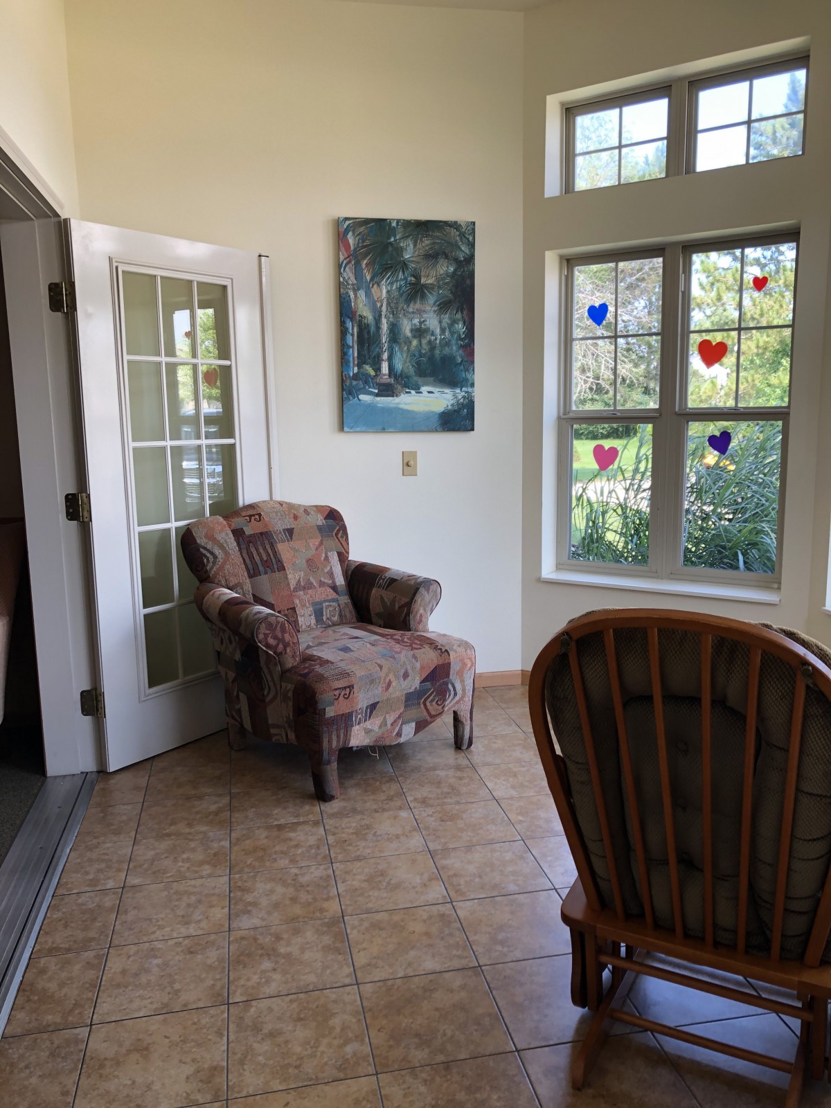 A cozy living room corner with a patterned armchair, art on the wall, and sunlight streaming through decorated windows.