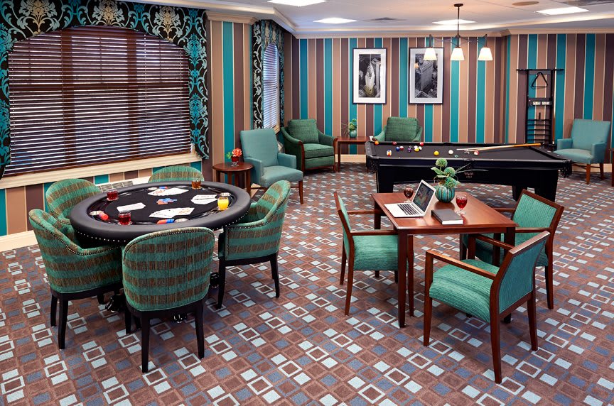 A stylish game room with a poker table and pool table, featuring striped walls, patterned carpet, and comfortable armchairs.